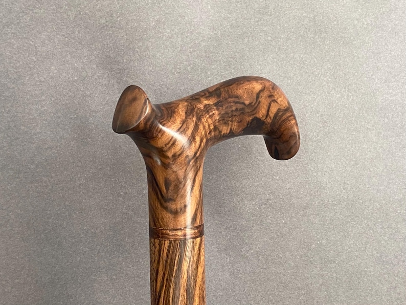 May include: A wooden walking cane with a curved handle. The cane is made of a dark brown wood with a unique grain pattern. The handle is smooth and rounded, providing a comfortable grip. The cane is a classic accessory.
