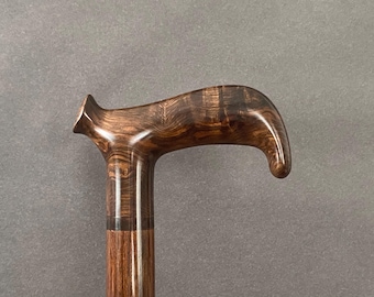 May include: A polished wooden cane handle featuring a dark brown finish with a prominent swirling grain pattern. The handle has a curved, ergonomic shape with a distinct protrusion at the top and a smooth, glossy surface texture throughout.