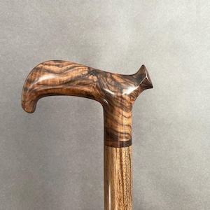 May include: A wooden walking cane with a curved handle. The handle is a dark brown with a unique wood grain pattern. The shaft is a lighter brown, and the cane appears to be made of multiple pieces of wood.