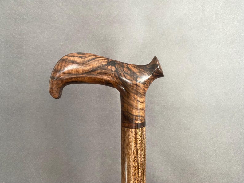 May include: A wooden walking cane with a curved handle. The handle is a dark brown with a unique wood grain pattern. The shaft is a lighter brown, and the cane appears to be made of multiple pieces of wood.