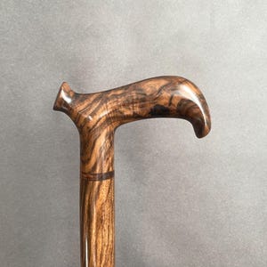 May include: A wooden walking cane with a curved handle. The cane is made of a dark brown wood with a prominent grain pattern. The handle is smooth and polished, and the shaft is cylindrical.