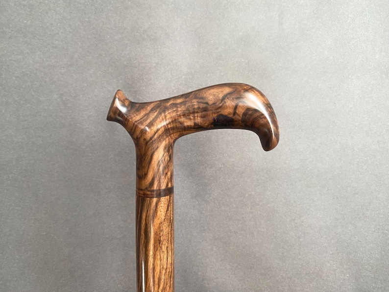May include: A wooden walking cane with a curved handle. The cane is made of a dark brown wood with a prominent grain pattern. The handle is smooth and polished, and the shaft is cylindrical.