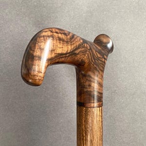 May include: A wooden walking cane with a curved handle. The handle features a rich, swirling grain pattern in shades of brown and tan. The shaft is a lighter brown, and the cane appears to be varnished for a glossy finish.