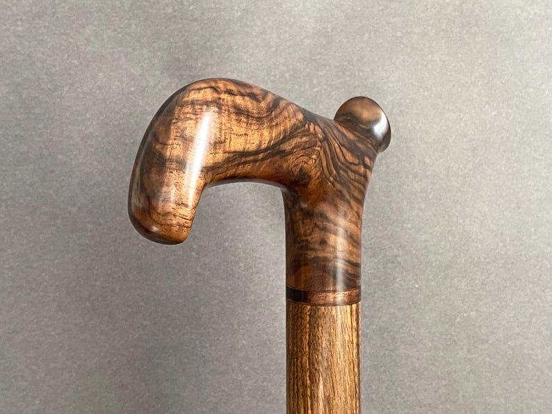 May include: A wooden walking cane with a curved handle. The handle features a rich, swirling grain pattern in shades of brown and tan. The shaft is a lighter brown, and the cane appears to be varnished for a glossy finish.