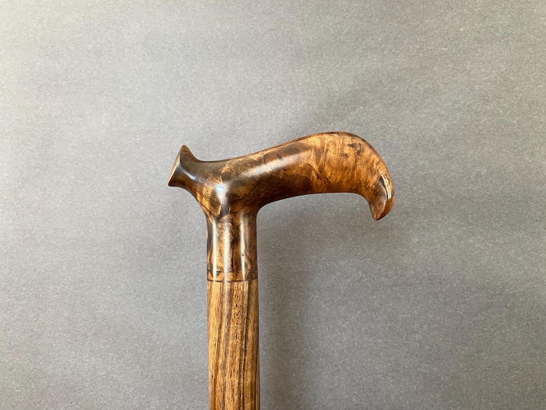 May include: A wooden walking stick with a curved handle. The handle is a dark brown with a polished finish, highlighting the wood grain. The shaft is a lighter brown, also with visible wood grain. The stick is designed for support and mobility.