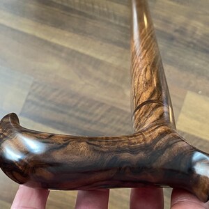 May include: A close-up of a wooden walking cane with a curved handle. The cane is made of dark brown wood with a prominent grain pattern. The handle is smooth and polished, with a rounded end for comfortable gripping.