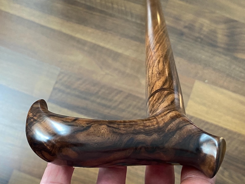 May include: A close-up of a wooden walking cane with a curved handle. The cane is made of dark brown wood with a prominent grain pattern. The handle is smooth and polished, with a rounded end for comfortable gripping.