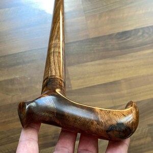 May include: A polished wooden walking stick with a curved handle. The stick is crafted from dark brown wood, displaying a prominent wood grain pattern. The handle is shaped for a comfortable grip and provides support for the user.