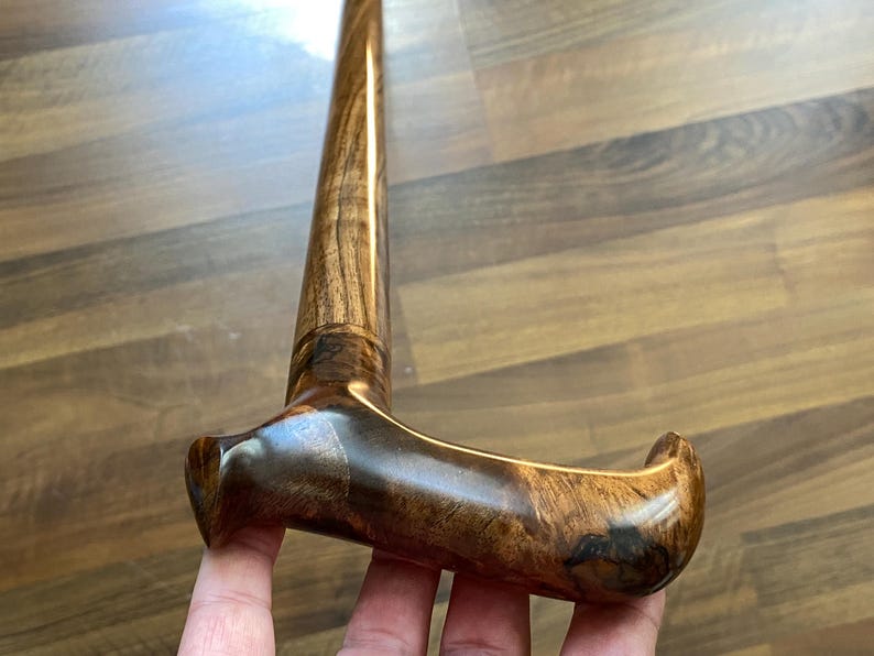 May include: A polished wooden walking stick with a curved handle. The stick is crafted from dark brown wood, displaying a prominent wood grain pattern. The handle is shaped for a comfortable grip and provides support for the user.