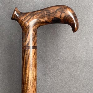May include: A wooden walking cane with a curved handle. The cane is made of a dark brown wood with a striped grain pattern. The handle is smooth and polished, and the cane appears to be sturdy and well-crafted.