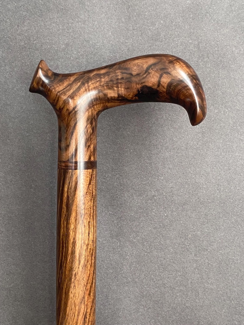 May include: A wooden walking cane with a curved handle. The cane is made of a dark brown wood with a striped grain pattern. The handle is smooth and polished, and the cane appears to be sturdy and well-crafted.