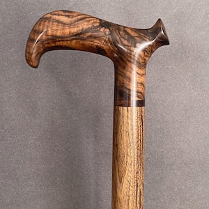 May include: A wooden walking cane with a curved handle. The handle features a dark, swirling grain pattern, while the shaft has a lighter, vertical grain. The cane is a rich brown color, with a polished finish.