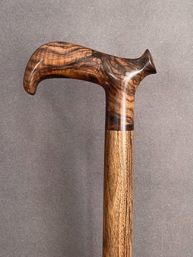 May include: A wooden walking cane with a curved handle. The handle features a dark, swirling grain pattern, while the shaft has a lighter, vertical grain. The cane is a rich brown color, with a polished finish.