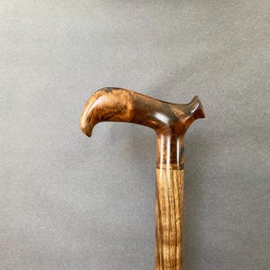 May include: A wooden walking cane with a curved handle. The handle is a dark brown with lighter wood grain. The shaft is a lighter brown with vertical grain. The cane is set against a grey background.