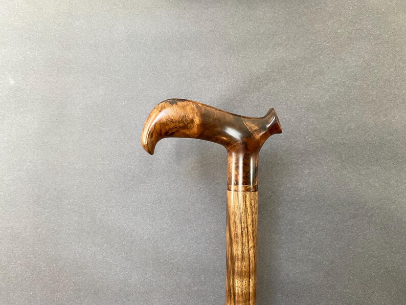May include: A wooden walking cane with a curved handle. The handle is a dark brown with lighter wood grain. The shaft is a lighter brown with vertical grain. The cane is set against a grey background.