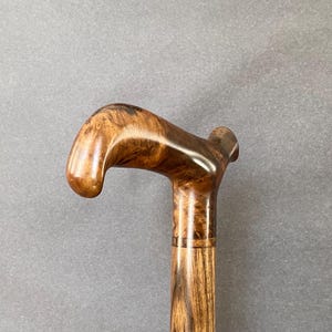 May include: A wooden walking stick with a curved handle. The handle and upper section are a glossy brown, while the shaft has a textured, lighter wood grain. The stick is designed for support and mobility, a traditional aid.