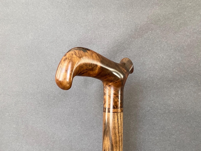 May include: A wooden walking stick with a curved handle. The handle and upper section are a glossy brown, while the shaft has a textured, lighter wood grain. The stick is designed for support and mobility, a traditional aid.