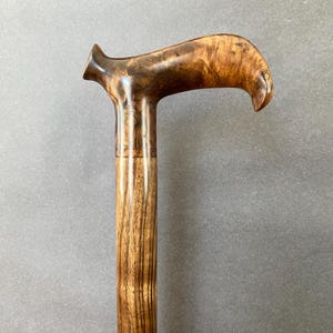 May include: A wooden walking stick with a curved handle. The handle is a dark brown with lighter wood grain. The shaft is a lighter brown with vertical grain. The cane is designed for support and mobility. The overall length is approximately 90 cm.