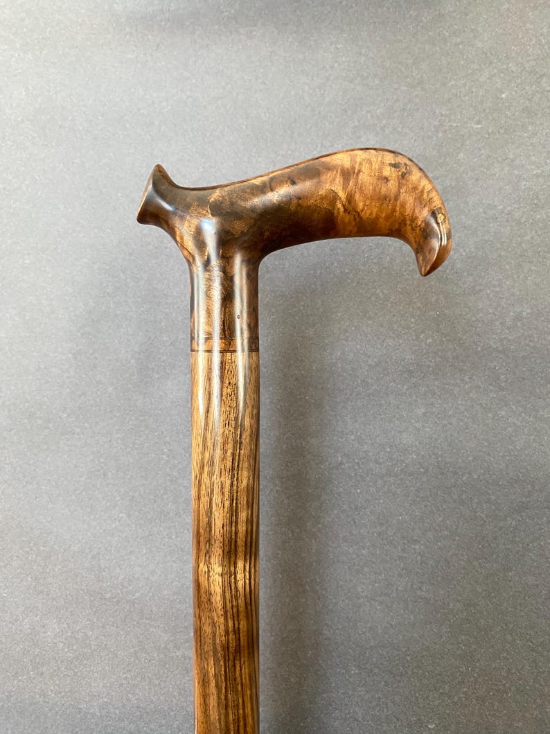 May include: A wooden walking stick with a curved handle. The handle is a dark brown with lighter wood grain. The shaft is a lighter brown with vertical grain. The cane is designed for support and mobility. The overall length is approximately 90 cm.