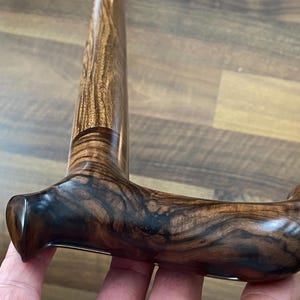May include: A close-up of a wooden walking cane with a curved handle. The handle and shaft are made of a dark brown wood with a prominent grain pattern. The handle is smooth and polished, with a curved design for comfortable gripping.