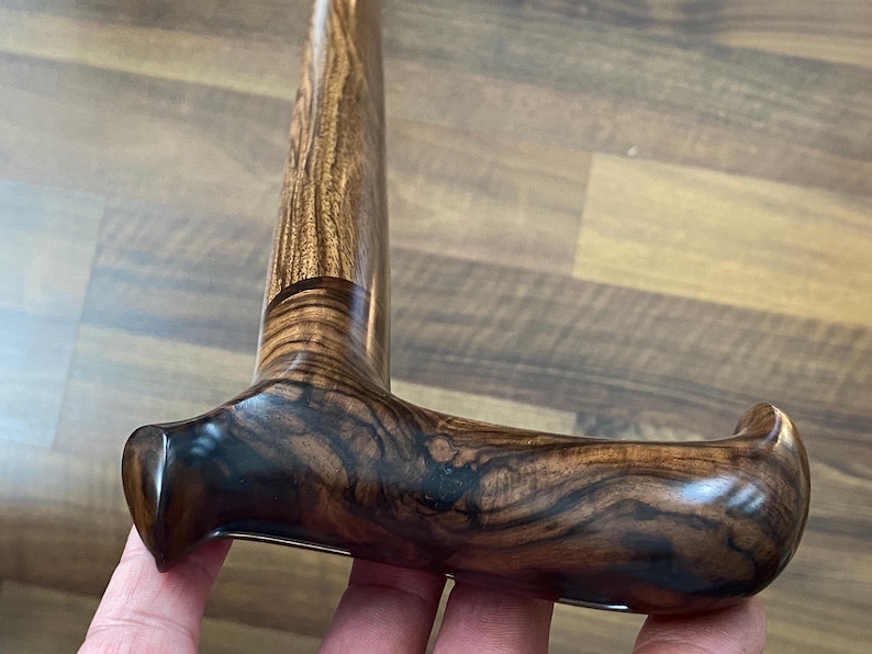 May include: A close-up of a wooden walking cane with a curved handle. The handle and shaft are made of a dark brown wood with a prominent grain pattern. The handle is smooth and polished, with a curved design for comfortable gripping.