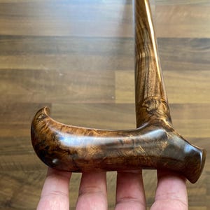 May include: A wooden walking stick with a curved handle. The cane is a deep brown colour, displaying detailed wood grain patterns. The handle is smooth and polished, highlighting the natural beauty of the wood. The cane is held by a person.
