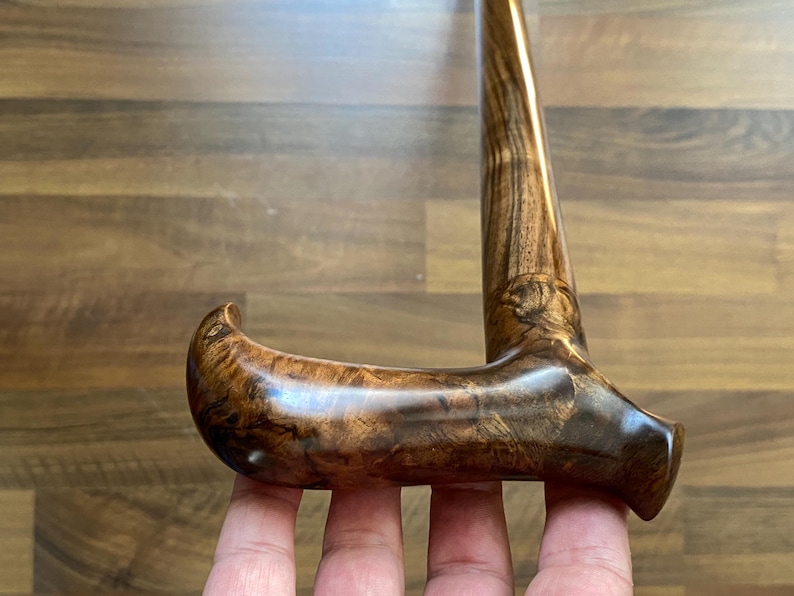 May include: A wooden walking stick with a curved handle. The cane is a deep brown colour, displaying detailed wood grain patterns. The handle is smooth and polished, highlighting the natural beauty of the wood. The cane is held by a person.