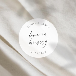 May include: A white circular sticker with the text "OLIVIA & JAMES" at the top, and "love is brewing" in cursive script in the center. The date "01.01.2026" is at the bottom. The sticker is on a white, textured background.