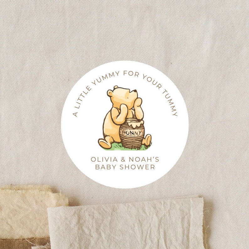 May include: A white round sticker with a cartoon Winnie the Pooh bear sitting on a green patch of grass holding a honey pot that says "Hunny". The sticker says "A little yummy for your tummy Olivia & Noah's Baby Shower".