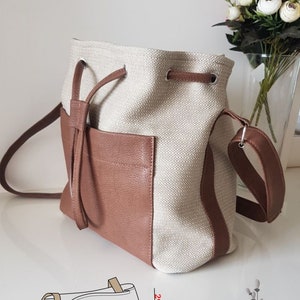 Könnte beinhalten: Eine beige und braune Eimer-Tasche mit Kordelzug und langem Trageriemen. Die Tasche ist 27 cm breit, 29 cm hoch und 14 cm tief. #modella_hervio