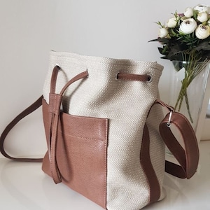 Peut inclure: Un sac seau en toile beige avec un rabat en cuir marron et une bandoulière en cuir marron. Le sac a une fermeture à cordon et une petite poche sur le devant.