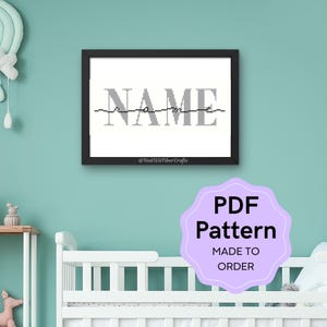 Op de afbeelding: Een ingelijst kruissteekpatroon met het woord "NAME" in grijze letters. Het patroon is beschikbaar als PDF-download en kan worden gepersonaliseerd met elke naam. De afbeelding toont het patroon in een kinderkameromgeving.