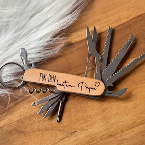 Könnte beinhalten: Ein Holz-Multitool mit verschiedenen Edelstahlwerkzeugen, darunter eine Schere und ein Korkenzieher. Das Werkzeug trägt die Gravur "FÜR DEN besten Papa" mit einem Herzsymbol. Das Werkzeug liegt auf einer Holzoberfläche.