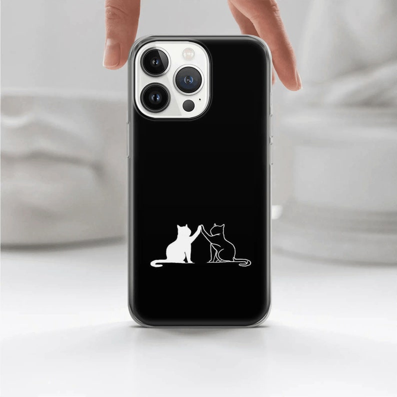 Puede incluir: Una funda de tel&eacute;fono negra con un contorno blanco de dos gatos d&aacute;ndose la mano. La funda del tel&eacute;fono tiene una parte posterior transparente y una parte delantera negra.