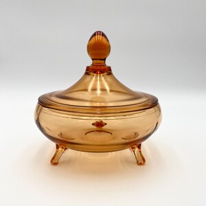 May include: A vintage amber glass candy dish with a lid and three legs. The dish has a rounded shape and a smooth, shiny finish.