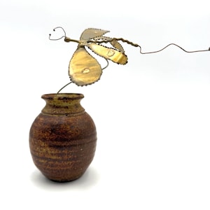 May include: A small, brown ceramic vase with a textured surface. A decorative metal dragonfly with gold-colored wings and a wire body extends from the vase. The dragonfly has a whimsical, handcrafted appearance.