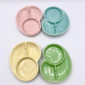 May include: Four ceramic serving dishes in pastel colors: pink, blue, yellow, and green. Each dish has a unique shape with a central circular compartment featuring playing card suit symbols: heart, spade, diamond, and club. The dishes have a speckled finish.