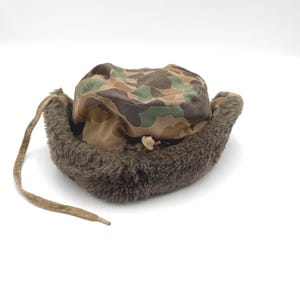 May include: A camouflage print trapper hat featuring a brown, green, and tan pattern on the crown. The hat includes a thick, dark brown faux fur lining along the brim and ear flaps, with a long, thin fabric tie hanging from the left side.