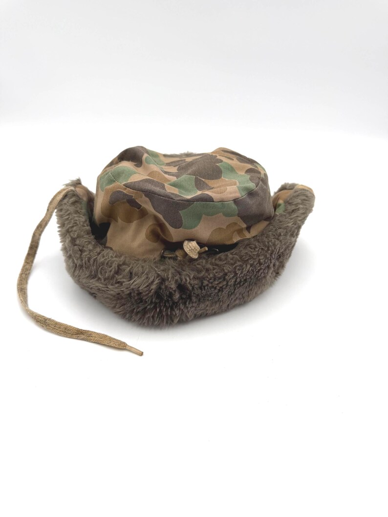 May include: A camouflage print trapper hat featuring a brown, green, and tan pattern on the crown. The hat includes a thick, dark brown faux fur lining along the brim and ear flaps, with a long, thin fabric tie hanging from the left side.