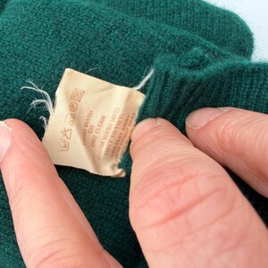 Puede incluir: Una prenda de punto verde oscuro con una etiqueta de cuidado beige que incluye instrucciones impresas para lavar a mano o en seco, y lavado a 40 grados Celsius. La etiqueta tambi&eacute;n muestra varios s&iacute;mbolos de cuidado para la prenda.