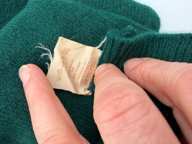 Puede incluir: Una prenda de punto verde oscuro con una etiqueta de cuidado beige que incluye instrucciones impresas para lavar a mano o en seco, y lavado a 40 grados Celsius. La etiqueta tambi&eacute;n muestra varios s&iacute;mbolos de cuidado para la prenda.