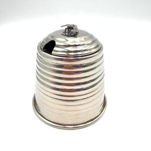 May include: A silver-colored honey pot with a bee-shaped handle on the lid. The pot has a cylindrical shape with horizontal ridges and a small opening for a spoon. The pot sits on a white surface.