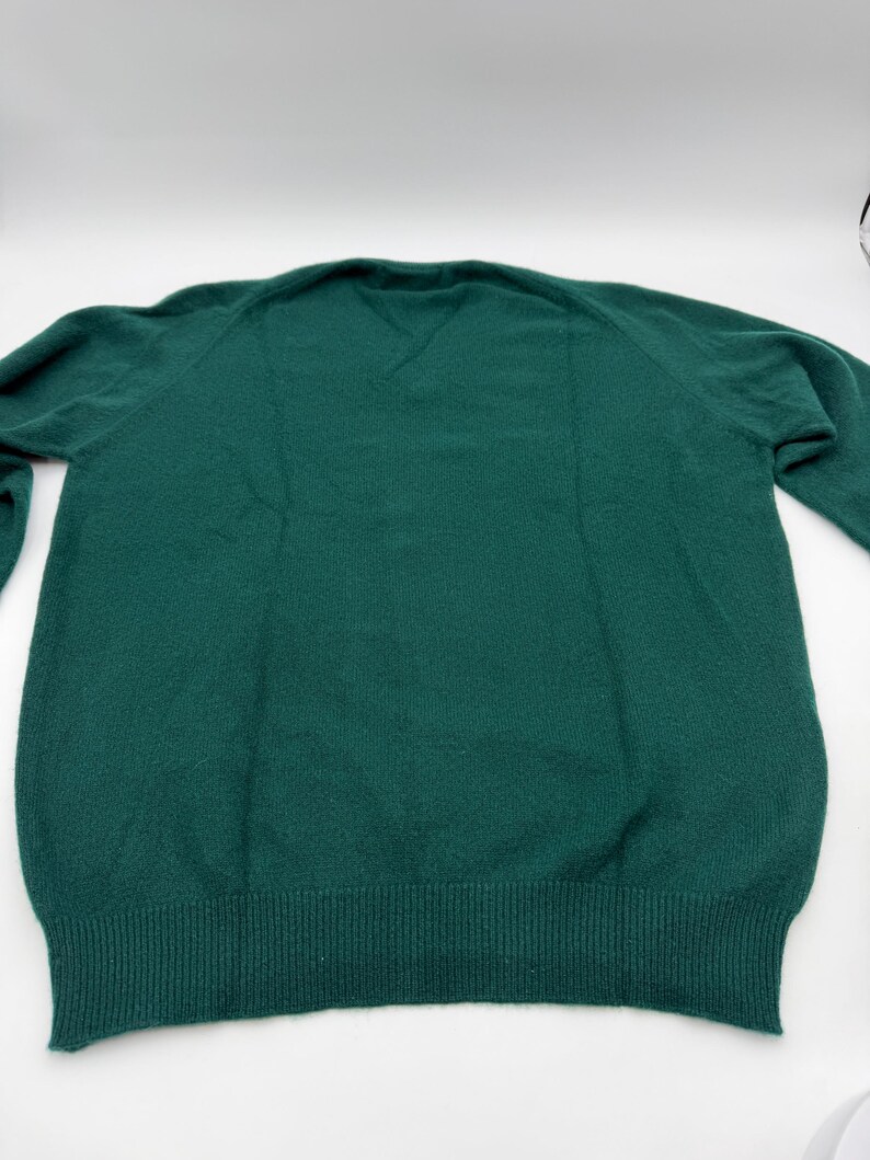 Puede incluir: Un su&eacute;ter de manga larga en color verde oscuro s&oacute;lido con cuello en V y pu&ntilde;os y dobladillo de punto acanalado. La prenda est&aacute; confeccionada con un tejido de punto suave y texturizado, con mangas ragl&aacute;n y un dise&ntilde;o limpio y sin estampados.