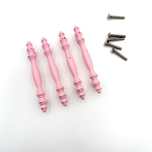 May include: Four light pink cabinet pulls with a distressed finish and decorative spindle-shaped detailing. Each pull features a slender, elongated form with rounded accents at the top and bottom. Four silver-toned metal screws are included.