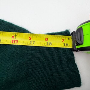 Puede incluir: Una manga de su&eacute;ter de punto verde oscuro con un pu&ntilde;o acanalado. Una cinta m&eacute;trica amarilla est&aacute; colocada sobre el pu&ntilde;o, mostrando un ancho de aproximadamente 18,5 cm. La tela tiene una apariencia suave y texturizada con puntos visibles.