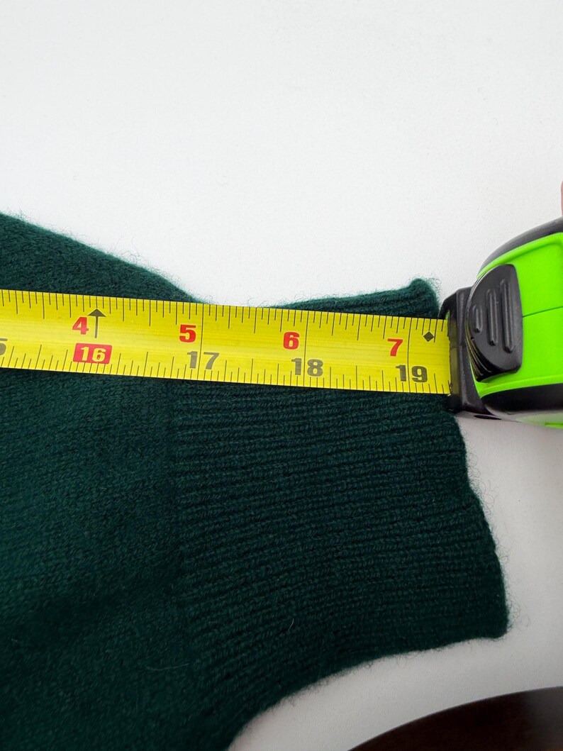 Puede incluir: Una manga de su&eacute;ter de punto verde oscuro con un pu&ntilde;o acanalado. Una cinta m&eacute;trica amarilla est&aacute; colocada sobre el pu&ntilde;o, mostrando un ancho de aproximadamente 18,5 cm. La tela tiene una apariencia suave y texturizada con puntos visibles.