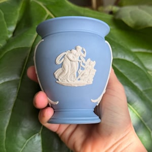 May include: A small, light blue vase with white relief figures. The vase has a rounded body and a flared rim. The relief depicts classical figures. The vase is held in a hand against a backdrop of green leaves.