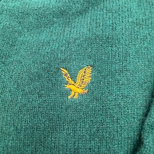 Puede incluir: Un su&eacute;ter de punto verde oscuro con una superficie de tela texturizada. Centrado en el pecho hay un logotipo de &aacute;guila amarillo dorado bordado, representado con las alas extendidas en posici&oacute;n de vuelo, mostrando costuras detalladas y un contraste vibrante.