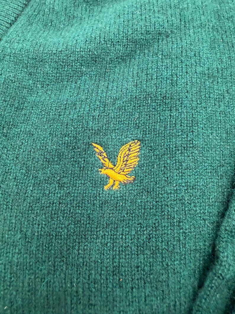 Puede incluir: Un su&eacute;ter de punto verde oscuro con una superficie de tela texturizada. Centrado en el pecho hay un logotipo de &aacute;guila amarillo dorado bordado, representado con las alas extendidas en posici&oacute;n de vuelo, mostrando costuras detalladas y un contraste vibrante.