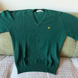 Puede incluir: Un su&eacute;ter de color verde oscuro con cuello en V, mangas largas y dobladillo y pu&ntilde;os acanalados. La prenda presenta un peque&ntilde;o logotipo de &aacute;guila amarilla bordado en el pecho izquierdo y una etiqueta interior blanca con el texto Lyle & Scott.